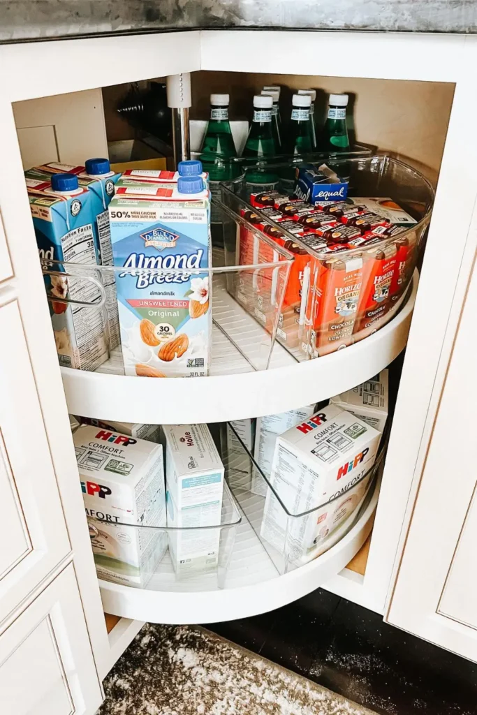 Corner pantry shelf with wooden lazy susan turntable holding jars of sauces, spices, and condiments, cozy warm kitchen lighting reflecting off glass jars, white pantry shelves with natural wood textures, woven baskets nearby, Pinterest-style organized kitchen corner with soft daylight and welcoming home atmosphere.