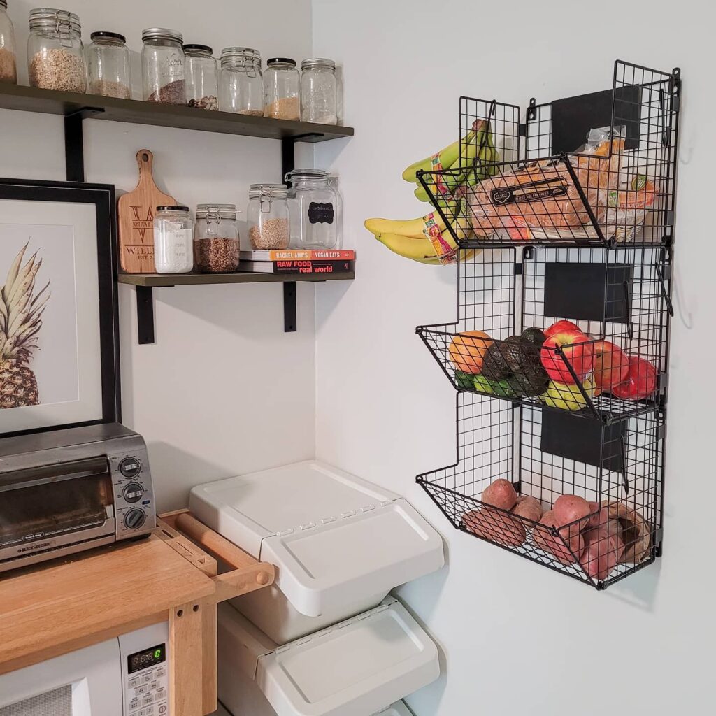 Pantry shelf with metal hanging baskets attached underneath, holding onions, garlic, and snack bags, warm cozy lighting highlighting textures of wooden shelves, modern farmhouse kitchen styling with neutral tones, Pinterest-style small pantry organization scene with woven baskets and soft natural daylight.